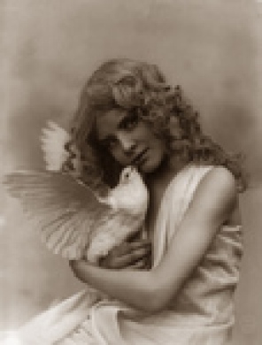 Sepia photo of a young caucasian girl with long curly hair, holding a white dove in her arms, 1902.